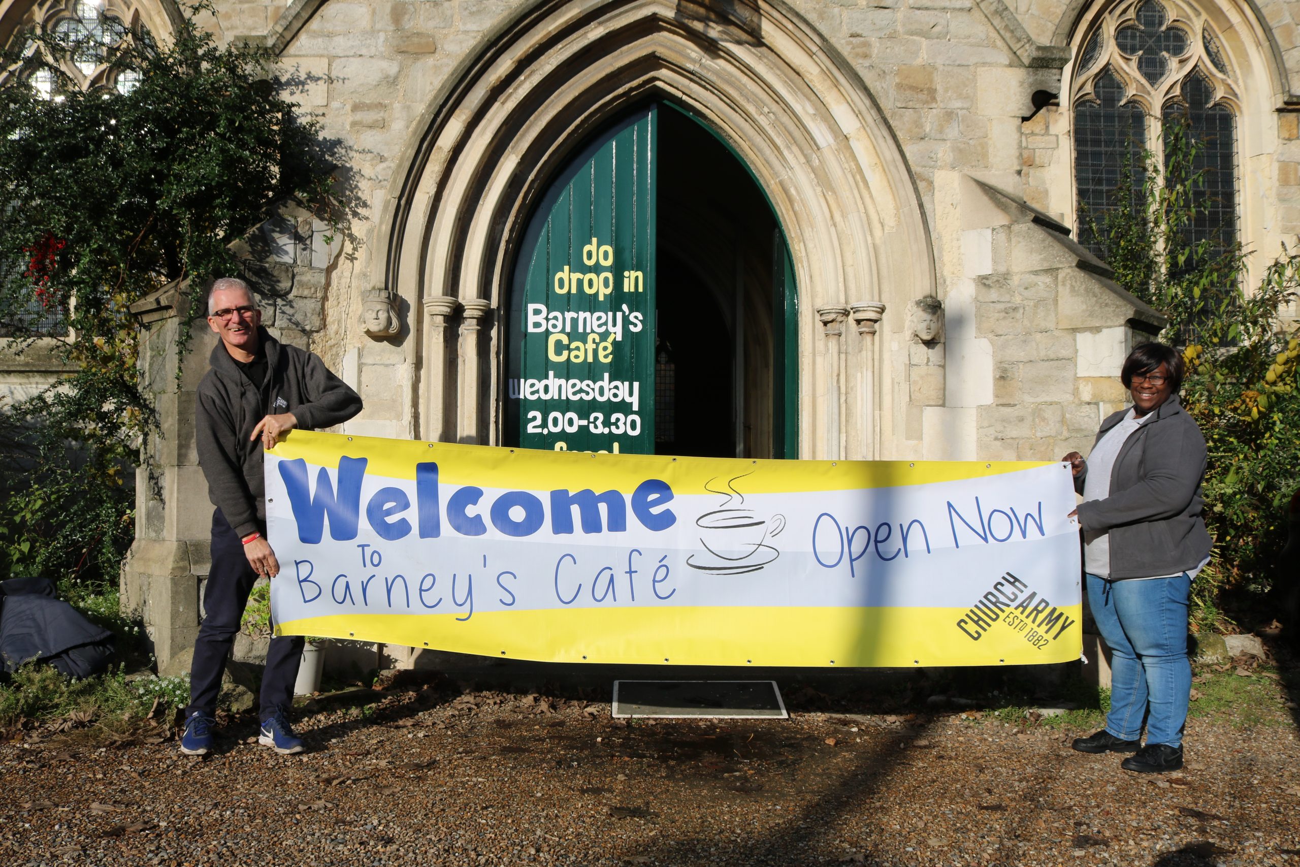 Stephen and Andrena holding a cafe welcome banner outside a church building
